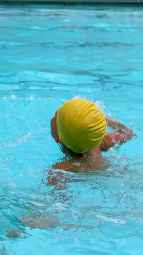 Vertical video: Camera starting filming child swimming practicing strokes at pool, yellow swim cap