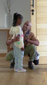 Returning father with gray duffel crouching to hug daughter in polka-dot sweater holding toy