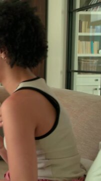 Vertical video: Turning woman in tank replying to offscreen voice on sofa, holding cup, copy space