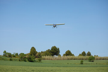 Fototapeta premium Small propeller airplane flying low above green rural field. Light aircraft in clear blue sky over peaceful countryside landscape.