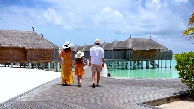 A happy family of four on summer vacations walks over a wooden jetty in the tropical, Maldives islands
