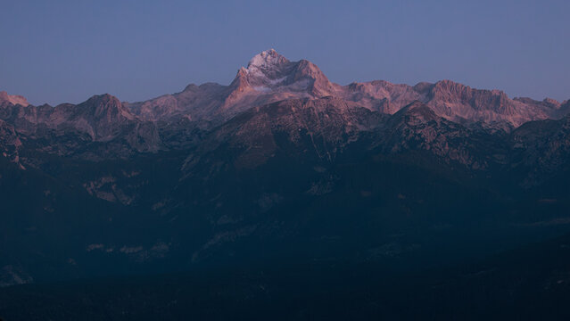 View of the majestic Triglav mountain peak glowing with pink alpine light at dawn over dark forested ridges Sorica, Skofja Loka, Slovenia.