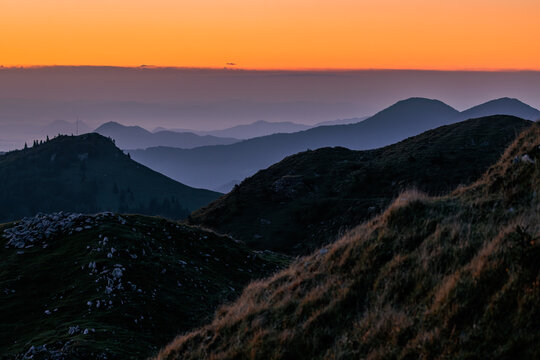 View of layered mountain silhouettes under a vibrant orange sunset sky with misty valleys in Sorica, Skofja Loka, Slovenia.