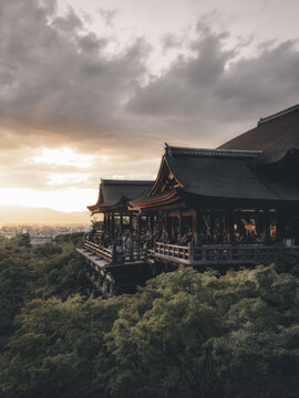 View of Kiyomizu-dera temple with a crowd of people on the wooden balcony overlooking lush green trees during sunset in Japan.