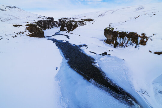 View of a river flowing through a snow-covered canyon with icicles on rocky cliffs under a blue sky in Iceland.