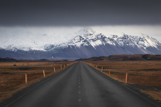 View of a straight asphalt road stretching toward massive snow-covered mountains under a heavy dark cloud layer in Iceland.
