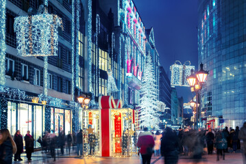 Obraz premium Crowded urban Christmas street scene at twilight: decorated city boulevard with cheerful pedestrians walking past storefronts