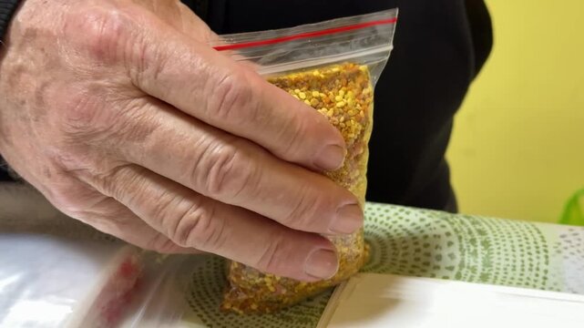 Close-up white male salesman packing bee pollen into transparent bag, hands pressing and sealing pouch on table with yellow
