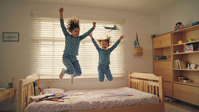 Two children in pajamas jumping with arms raised on bed laughing joyfully in bright morning light in a colorful toy-filled bedroom. Childhood happiness concept