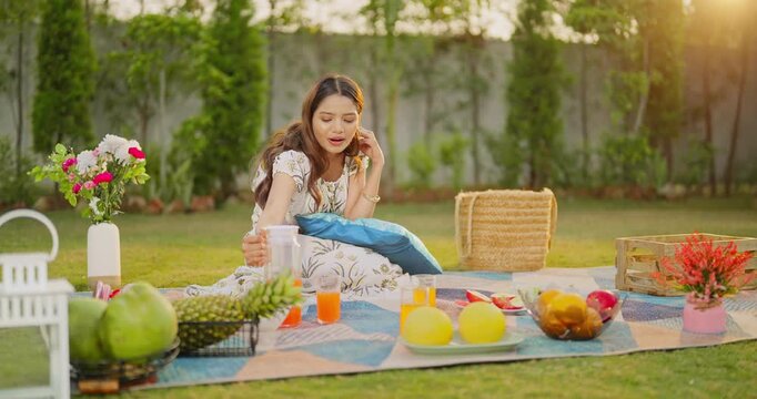 Indian young adult gen z female sitting on mat pick up jug pour fresh orange juice into glass enjoy new picnic trip place happy teen girl play fruits spend good day time do self care have fun joy