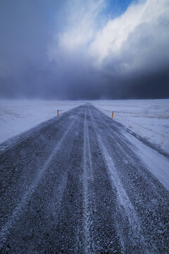 View of a straight asphalt road covered in snow and ice stretching through a vast winter landscape under a dramatic cloudy sky in Snaefellsnes, Eyja- og Miklaholtshreppur, Iceland.