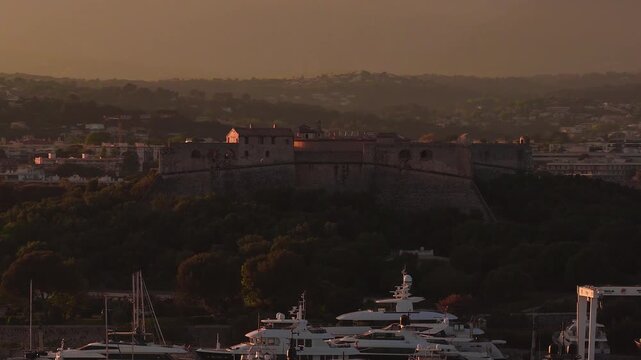 Aerial drone pan sweeps Antibes, France at golden hour. Fort Carre crowns a wooded hill above Port Vauban as superyachts rest in the marina, warm light casts long shadows.