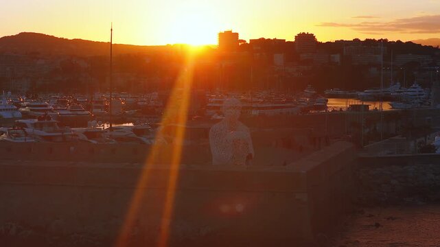 Aerial glide over Antibes, France shows Port Vauban, Le Nomade, moored yachts, golden light, lens flares, silhouetted skyline, reflective hulls, calm mood.