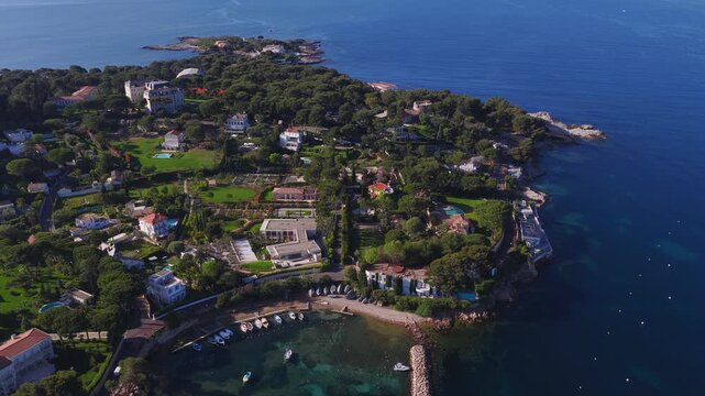 Aerial bird's eye view of Cap d'Antibes, French Riviera, with pine groves, villas, a chateau like estate, gardens, pebble beach, breakwater, moored boats, and turquoise waters