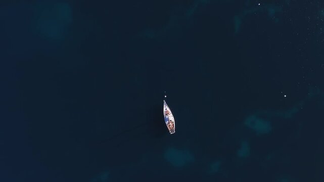 Aerial view shows a lone sailboat anchored off Antibes, France, gently drifting near white buoys over navy water and turquoise seabed patches at sunset.