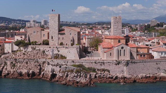 Aerial view of Antibes on the French Riviera shows a medieval stone fortress with two square towers, old town roofs, plaza visitors, and calm waves under clear skies.