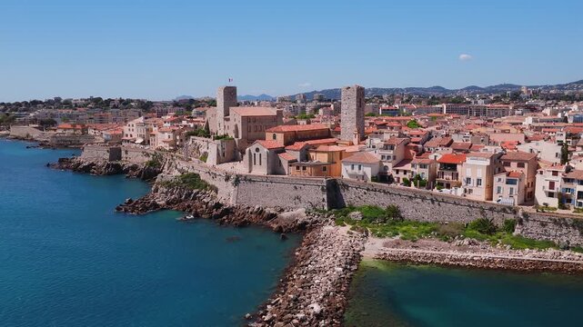 Aerial view of Antibes old town, Chateau Grimaldi, two towers and flag. Camera glides over turquoise sea, breakwater, terracotta roofs, seawall walkers at midday.