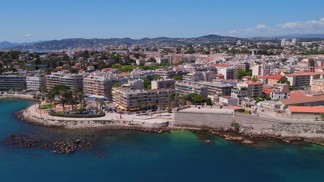 Aerial view of Antibes, France shows rocky shore, clear sea, old town ramparts, a bastion wall, palm promenade, cafes, terracotta roofs, and hills in midday light.