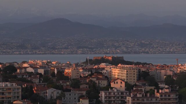 Aerial sunset panorama of Antibes, France, shows Fort Carre, terracotta rooftops, low rise blocks, yacht masts, calm bay, distant coastline, hills, and mountains.