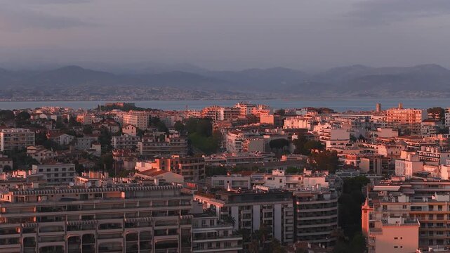 Aerial view of Antibes, France at sunset shows Fort Carre, a clock tower, apartment blocks, terraces, narrow streets, coastline, and hazy hills by the sea.