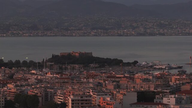Aerial dusk view over Antibes on the French Riviera shows Fort Carre, terracotta roofs, marina masts and yachts, lighthouse, breakwater, calm water, distant hills.