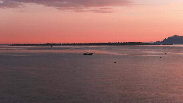 Aerial sunset over the Mediterranean at Antibes shows tree lined headlands, Esterel like peaks, a two masted yacht, buoys, faint tender wake, and gentle ripples.