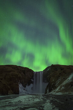 View of Skogafoss waterfall with the green Aurora Borealis dancing in the starry night sky over dark cliffs in Vik, Myrdalshreppur, Iceland.