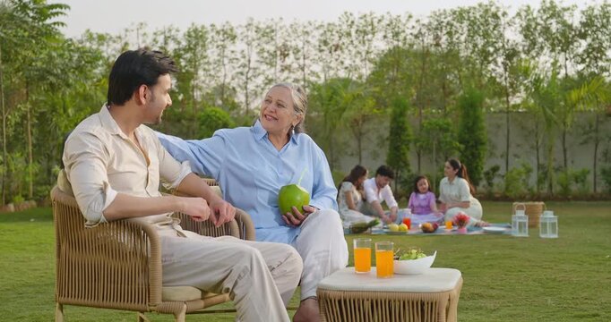 Indian old aged mom sitting on chair with young adult son talk share gossip hold fresh coconut drink juice outdoor Happy male man older female speak funny spend time enjoy picnic trip day garden place