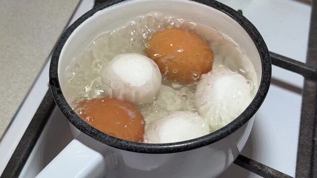 Close-up boiling eggs in enameled ladle on gas stove, brown and white chicken eggs simmering in bubbling water, steam rising, white