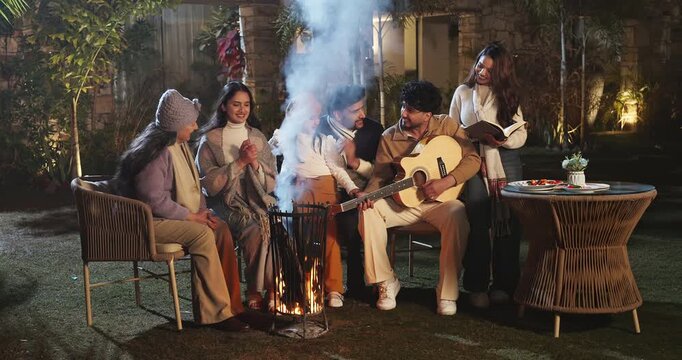 Indian mature adult female male sitting on chair man hold guitar play music sing song enjoy cold season happy family group girl child kid clap hand spend night time have fun joy clap hand garden place