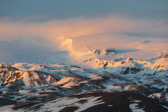 View of snowy mountain peaks illuminated by golden sunset light with soft clouds rolling over the ridges in Vik, Myrdalshreppur, Iceland.
