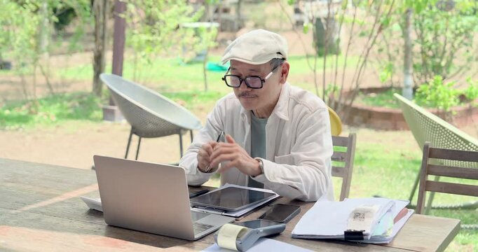 Asian father reviews laptop data, tablet, payment terminal, paperwork, coffee cup at garden cafe table, planning family restaurant strategy, accounting, stock workflow, service standard guide.