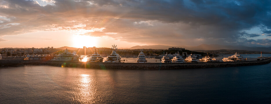 Panoramic aerial view of Antibes, France, shows superyachts at Port Vauban as the sun sets behind low mountains, old town bastions on the right, calm harbor water glinting.