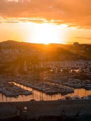 Fototapeta premium Warm sunset bathes Antibes old harbor and stone seawall, yachts and sailboats rest in calm water, masts form lines, backlit hills and silhouetted townscape add depth.