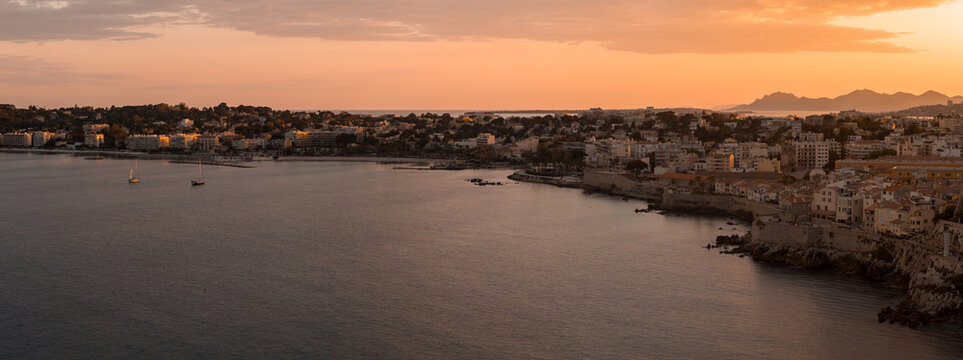 Aerial view of Antibes, France at sunset. Old town stone buildings line the rocky shore, calm Mediterranean in front, Fort Carre and ramparts visible, boats on the horizon.