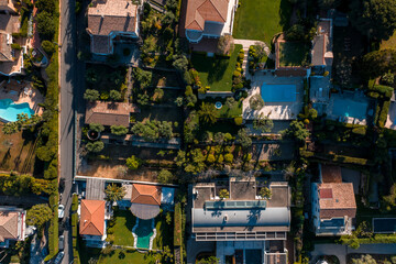 Aerial view of Antibes, France, with villas, terracotta roofs, turquoise pools, hedges, and tree lined paths at golden hour. A modern glass roofed villa stands out. © Aerial Film Studio