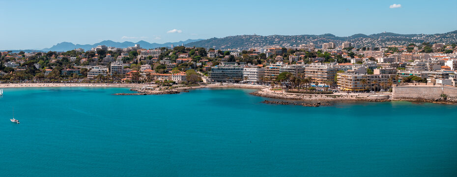 Aerial view of Antibes, France, shows Baie des Anges, sandy beaches, a marina, and Vieil Antibes ramparts. Late afternoon light, calm water, lone sailboat, Alps backdrop.