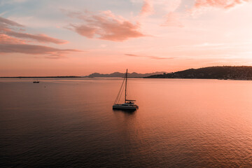 Aerial view of a solitary sailboat drifting on calm rose gold waters near Antibes, France at sunset, pastel sky reflecting across the bay with dusky hills beyond. © Aerial Film Studio