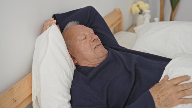 Grey haired senior man lying on bed, eyes closed, hand resting on chest and arm bent behind head in a residential building bedroom by wooden headboard and vase; serenity.