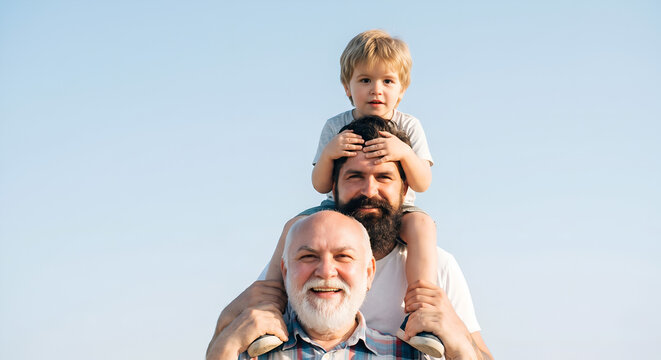 Three generations of smiling men with a grandfather carrying his adult son and young grandson on his shoulders against a clear blue sky