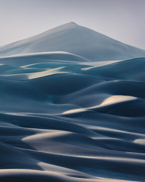 View of rolling sand dunes with soft blue shadows and a prominent triangular peak under a hazy sky in Balochistan, Pakistan.