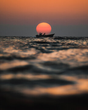 View of a small boat with people silhouetted against a large setting sun over the calm sea waves in Balochistan, Pakistan.