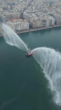 THESSALONIKI, GREECE - JANUARY 6, 2024: Vertical aerial drone shot orbiting and ascending over a fireboat spraying water cannons in the Thermaikos Gulf, revealing the commercial port of Thessaloniki d