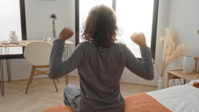 Man on bed sits at edge and stretches both arms upward toward window with desk and pampas grass in bedroom; morning calm.