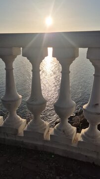 Sunset ocean balustrade viewed through white stone railings at the Mediterranean coast during golden hour with bright sun flares and calm water