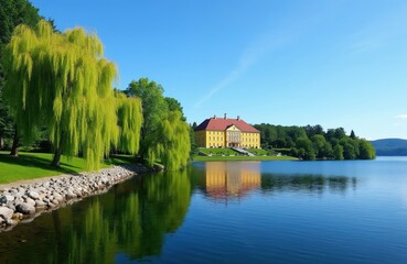 Obraz premium Yellow mansion with red roof sits on green grass by calm blue lake. Weeping willow trees fringe grassy shore, reflecting in water under clear sky.