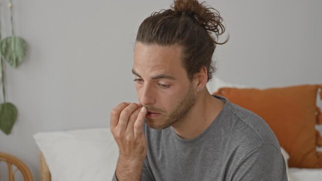 Man bites fingernails with hand to mouth on bed in bedroom, seated and slightly tense; anxiety introspection.