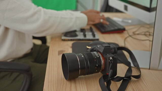 Young man photographer with mirrorless camera and lens on desk, hand typing on keyboard in studio; creative focus.
