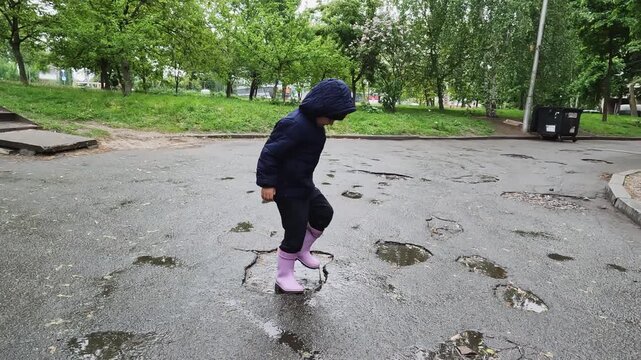 A small child wearing a jacket and boots stands in a muddy puddle on cracked asphalt. Keywords: childhood, weather, rain, exploration, discovery, learning, adventure, mud, curiosity, ecology, city