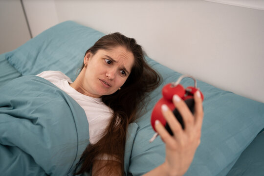 Bad morning concept. Shocked overslept woman waking up at home holding alarm clock lying in bed late for work . Oversleeping Problem.